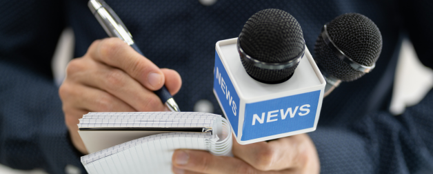 close up image of two hands holding a "news" microphone and writing notes on a small notepad