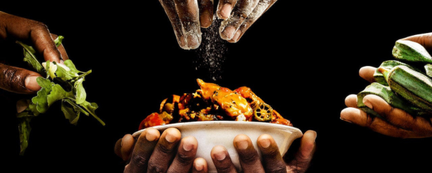 two hands holding a bowl of food and three other sets of hands contributing to the bowl with different ingredients of okra, spices, and greens.
