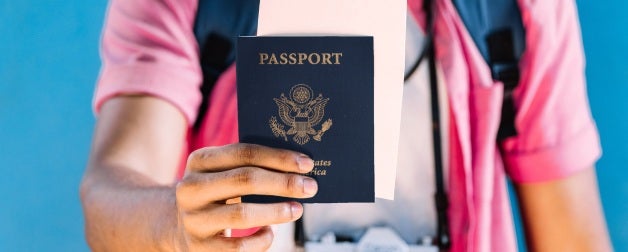 Person wearing white shirt and pink open button down with backpack and camera around neck holding US Passport 