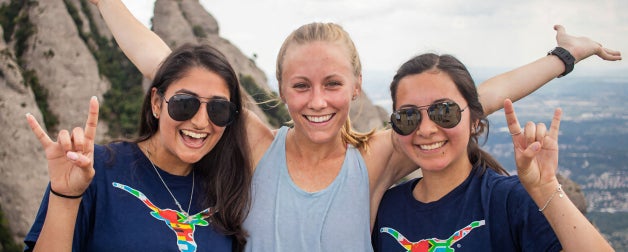 Three smiling female students abroad display the hook'em sign on a high peak