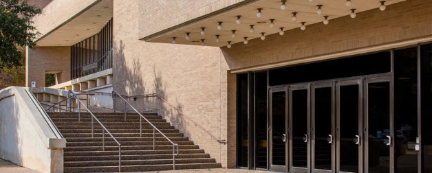 Building entrance with multiple glass doors and a large overhead marquee. Adjacent stairs with metal railing lead to an upper walkway.
