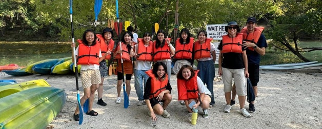 Smiling ELC students wearing lifejackets with paddles and kayaks pose in front of Barton Springs