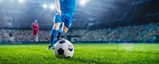 Soccer player runs towards the ball on grass field of a stadium, backdropped by lights and a seats full of cheering fans