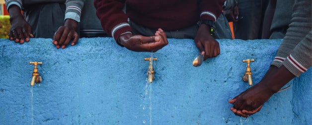 Tanzanian hands scooping and drinking water from brass fountains set against a light blue stone wall