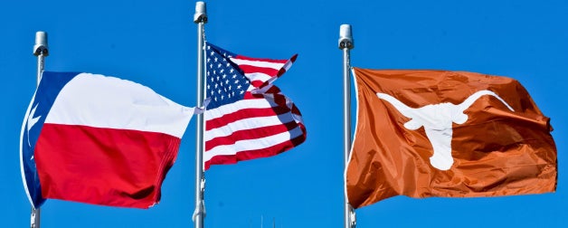 Texas state flag, American flag, and University of Texas Longhorns flag waving against a clear blue sky.