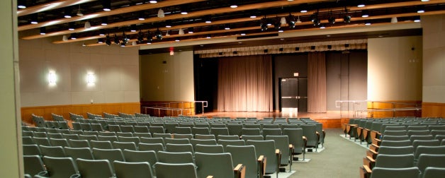 Empty auditorium with rows of gray seats facing a stage with closed curtains and overhead lighting.