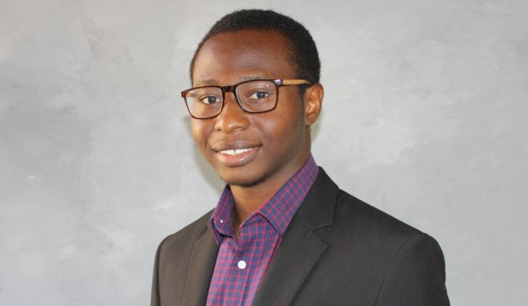A researcher from Nigeria in a suit poses for a portrait in front of a gray backdrop.