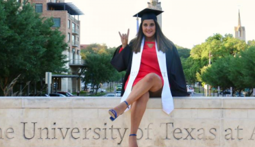 Marielisa sits on UT-Austin sign with her cap and gown and a hook 'em horns hand sign 