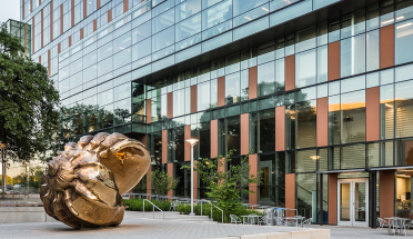Dell Medical School's windowed exterior reflects the sunset light. A golden conch seashell sculpture commands the foreground. 
