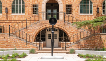 Sentinel IV, a bronze sculpture with a thin, long body with a large half-sphere head, stands in the middle of the Anna Hiss Gymnasium Courtyard on a bright day.
