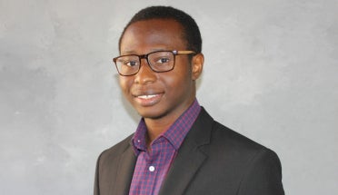 A researcher from Nigeria in a suit poses for a portrait in front of a gray backdrop.