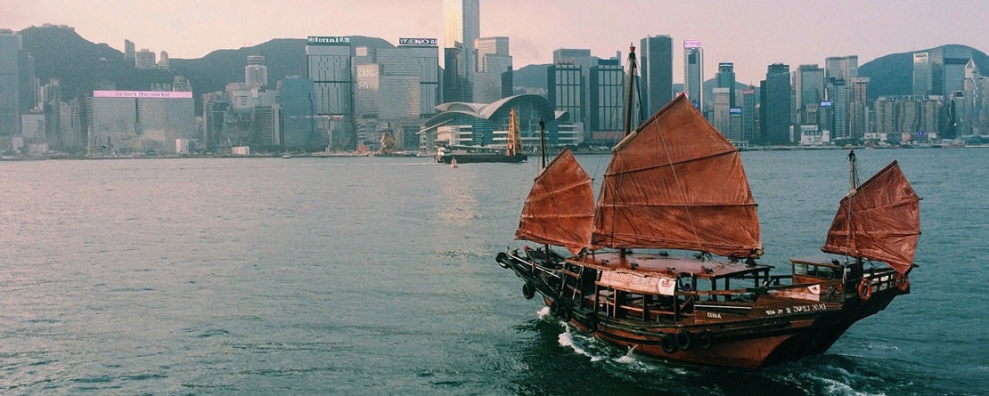 An asian sailboat sails toward the city on calm ocean waters amidst a backdrop of a pink sunset