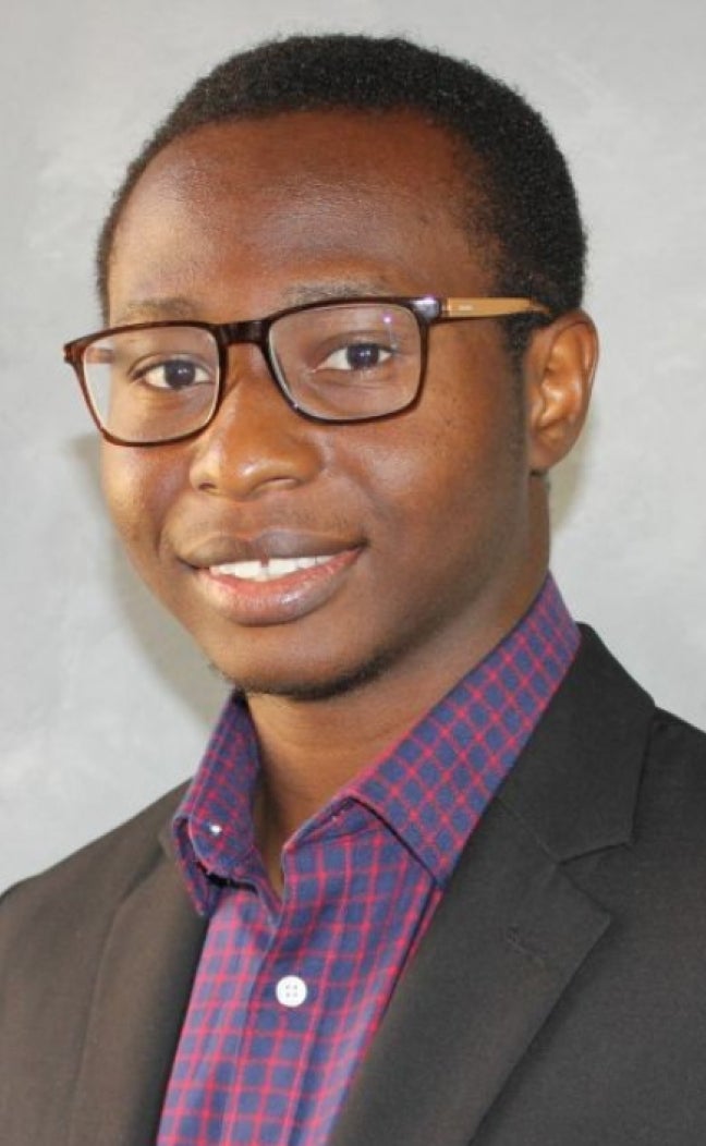 A researcher from Nigeria in a suit poses for a portrait in front of a gray backdrop.