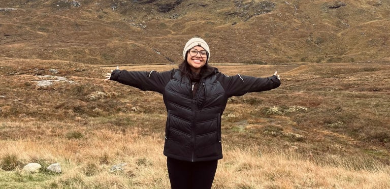 Elizabeth stands with her arms open and big smile backdropped by tall, rocky mountains