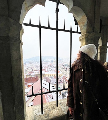 Jackie Villalobos looks out on a Europen city from a medieval-looking window