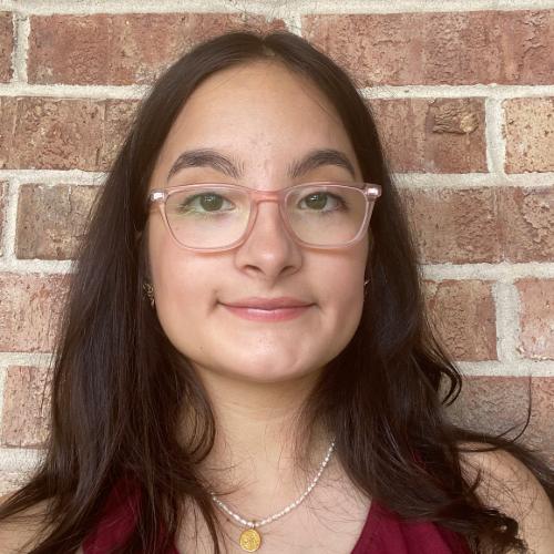 student in a red top smiles in front of a brick wall. student is wearing glasses.