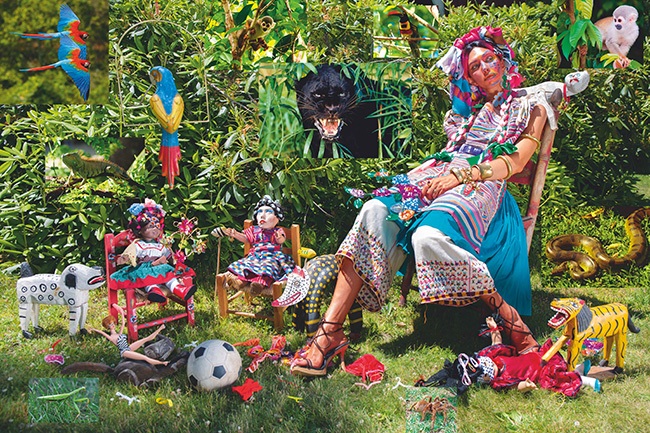 A medium dark-skinned woman in a colorful, intricately patterned outfit surrounded by dolls and other toys in a garden with images of jungle animals in the background