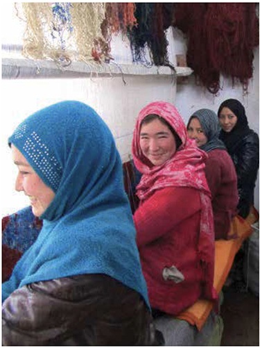 Four women in headscarves smile as they weave a rug on a loom