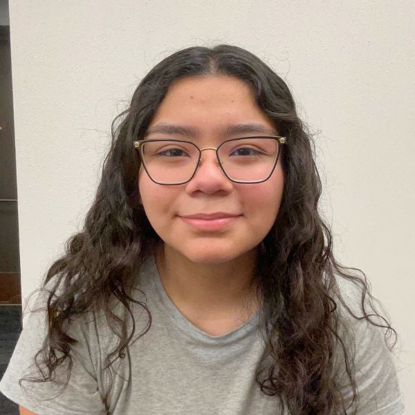 Young person with long, curly hair and glasses, smiling softly against a plain background. They wear a gray shirt, creating a calm and approachable tone.