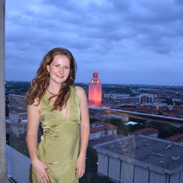 A woman in a green dress smiles on a rooftop, with a cityscape featuring a tall, illuminated tower in the background under a cloudy, evening sky.