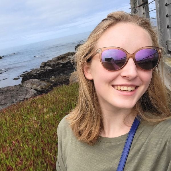 Smiling person with sunglasses takes a selfie near a rocky coastline. Overcast sky and ocean in the background suggest a serene, joyful mood.