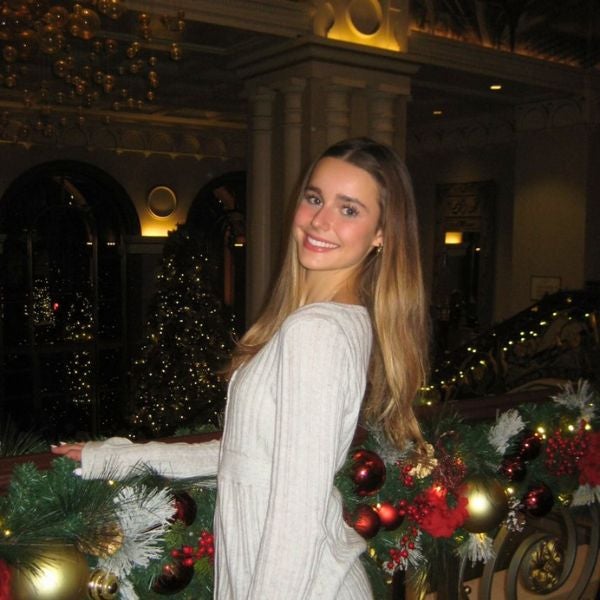 A woman in a light grey dress smiles while standing beside a decorated railing with festive red and gold ornaments. The background has sparkling Christmas trees, creating a joyful holiday atmosphere.