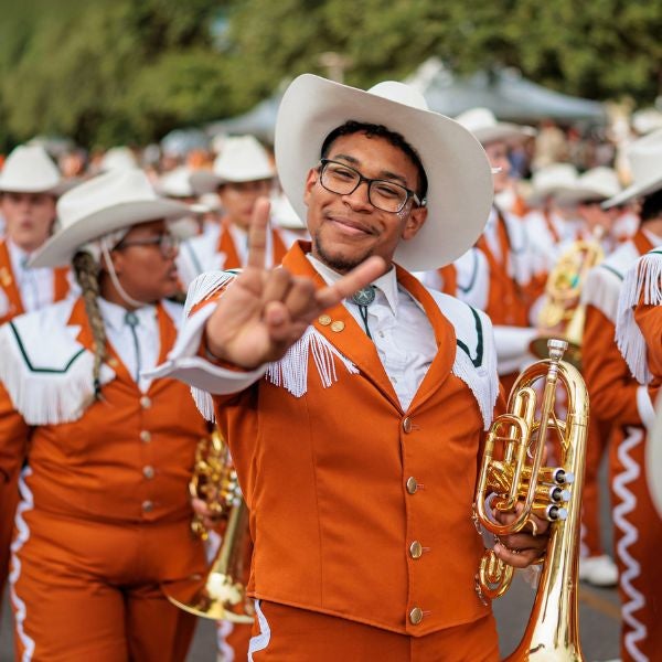 A joyful marching band member in orange and white uniform holds a brass instrument, smiling and making a hand gesture. The band is in festive, high spirits.
