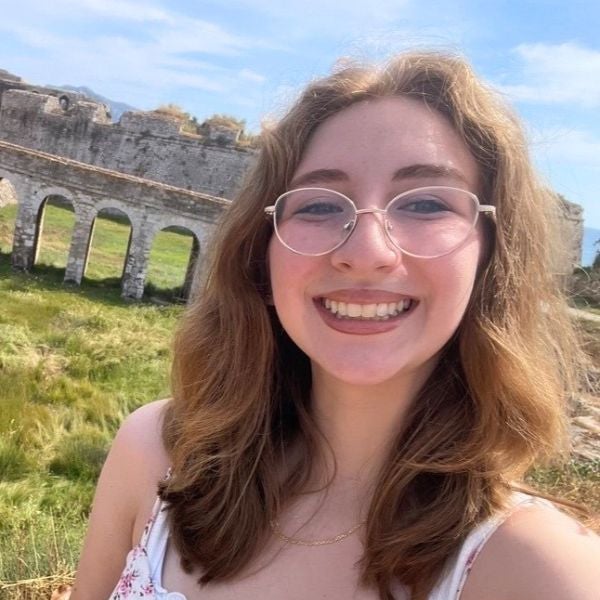 A young person with glasses smiles in front of ancient stone arches. The scene is sunny with green grass and a blue sky, conveying joy and exploration.