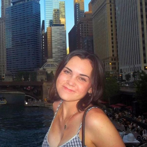 A person smiling at the camera with a cityscape background featuring tall buildings and a river. The scene conveys a warm, relaxed urban atmosphere.