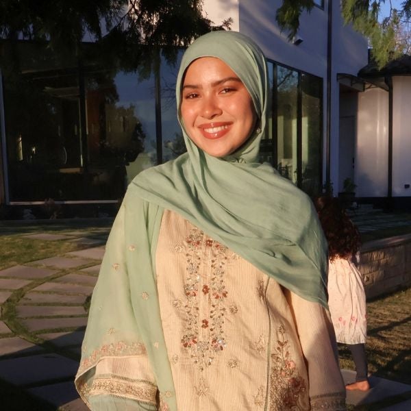 Smiling woman in a mint green hijab and embroidered beige outfit, standing in sunlight in a garden with a modern house in the background.