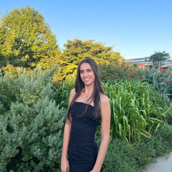 A woman in a black dress stands smiling in a lush garden. Greenery and blooming plants surround her under a clear blue sky, creating a serene atmosphere.