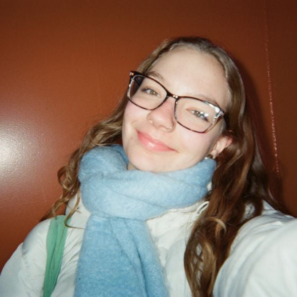 Smiling person with glasses and long hair, wrapped in a blue scarf, against a warm brown background. The mood is friendly and cheerful.