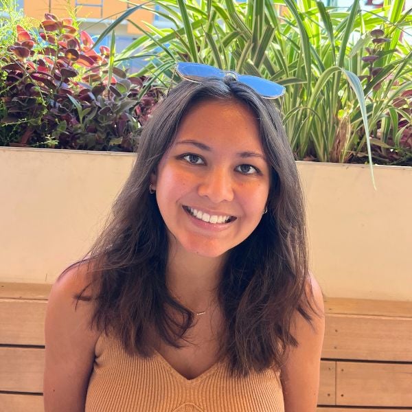 Smiling woman with long hair and sunglasses on her head, sitting on a bench. Lush green plants in the background, creating a bright, cheerful vibe.
