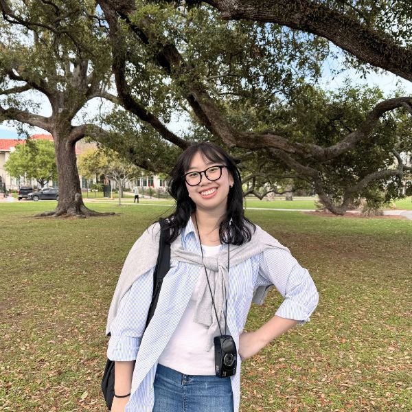Smiling person with glasses stands in a park, wearing a striped shirt and jeans. A camera hangs from their neck. Large trees in the background.