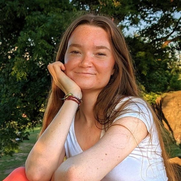 Young woman with long brown hair and a serene smile sits outdoors. She rests her chin on her hand. Sunlight illuminates her face and green foliage.