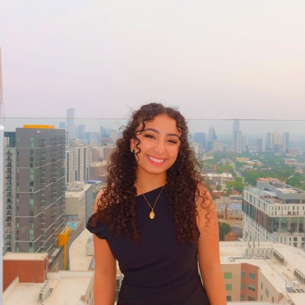 Smiling woman with curly hair in a black dress stands on a rooftop, with a cityscape in the background under a hazy, overcast sky.