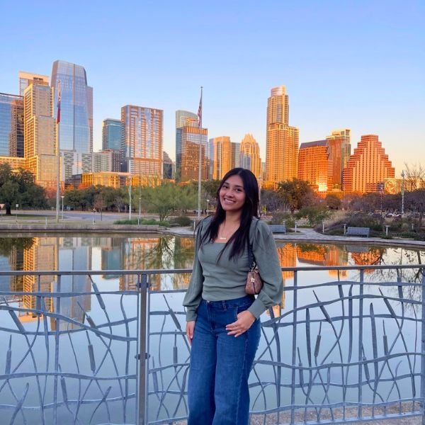 Smiling woman stands on a bridge with intricate railing, city skyline reflecting in water at sunset behind her, creating a warm, serene atmosphere.