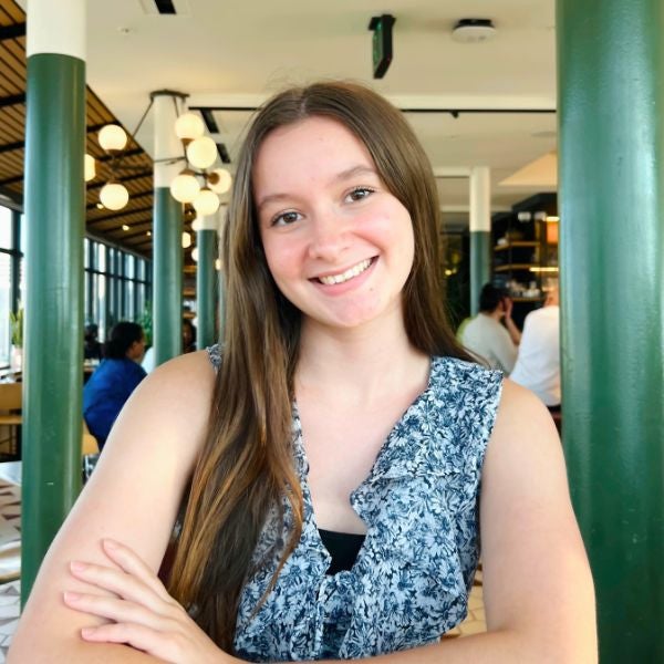 A young woman with long brown hair smiles warmly, seated in a stylish restaurant with globe lights and green columns. The atmosphere is bright and inviting.