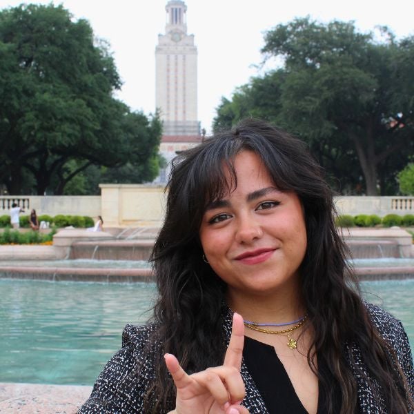 Smiling woman in a black blouse and layered necklaces makes a "hook 'em" hand gesture. She stands in front of a fountain and university tower, surrounded by trees.
