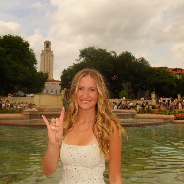 A smiling woman in a white polka-dot dress stands by a campus fountain, making a gesture with her hand. Tower and trees are visible in the background.