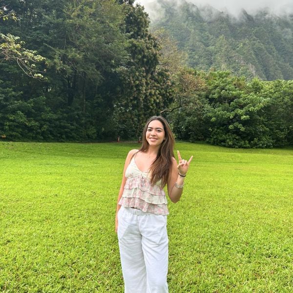 A woman stands on a vibrant green lawn, making a hand gesture. She's wearing a white, layered top and pants. Lush trees and misty mountains are in the background.