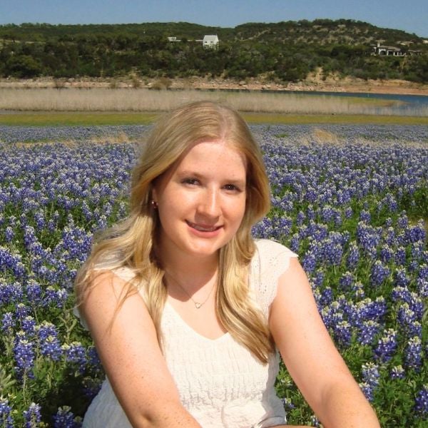 Young woman with long blonde hair and a white dress sits in a vibrant bluebonnet field, smiling contentedly. A serene landscape is visible in the background.