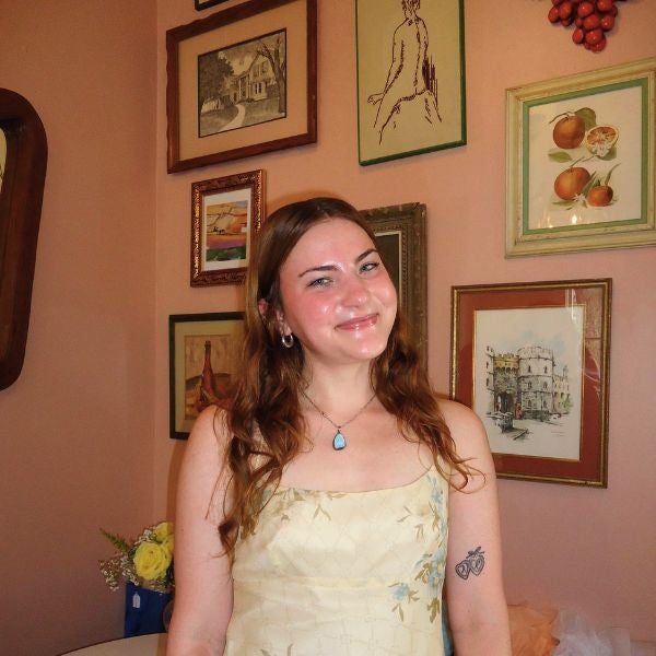 Smiling woman in a light dress stands in front of a wall adorned with framed artwork, exuding warmth and positivity. A vase of flowers is visible in the background.
