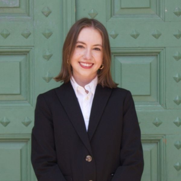 Smiling woman in a black blazer and white shirt stands against a textured, green door. The professional setting conveys confidence and warmth.