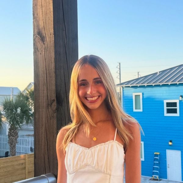 Smiling young woman in a white top stands outdoors at sunset, with a blue building and palm tree in the background, conveying a joyful, relaxed vibe.