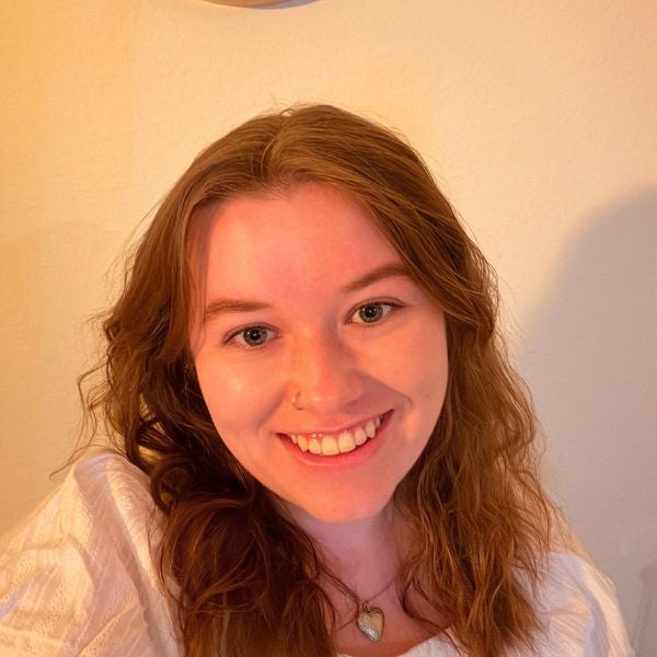 Smiling woman with wavy brown hair, wearing a white top and heart necklace. Warm lighting creates a cheerful and friendly atmosphere.