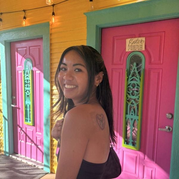 Smiling woman with long hair stands in front of vibrant pink doors with teal trim and yellow walls, displaying a heart tattoo on her shoulder.