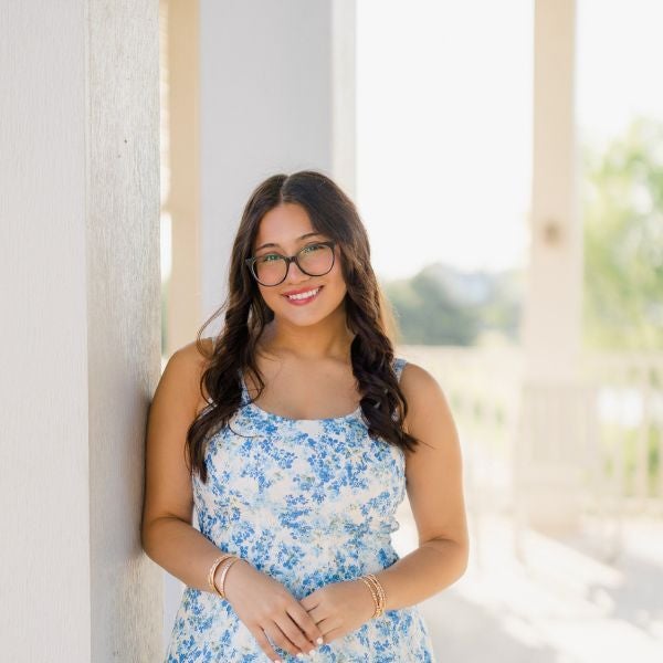 Woman with wavy hair and glasses, smiling in a blue floral dress, leans against a pillar on a sunny porch, conveying a cheerful and relaxed vibe.