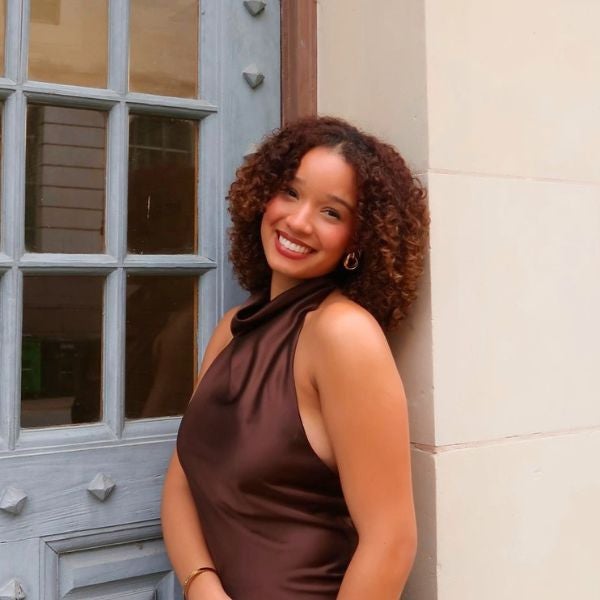 Woman with curly hair wearing a sleeveless brown dress leaning against a beige wall near a gray door with decorative studs.