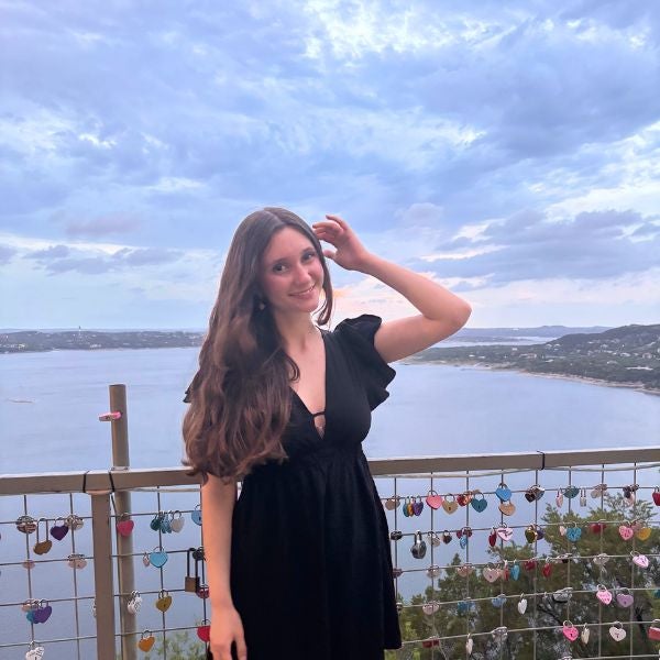 Young woman in a black dress stands by a railing adorned with colorful love locks overlooking a serene lake under a cloudy sky.